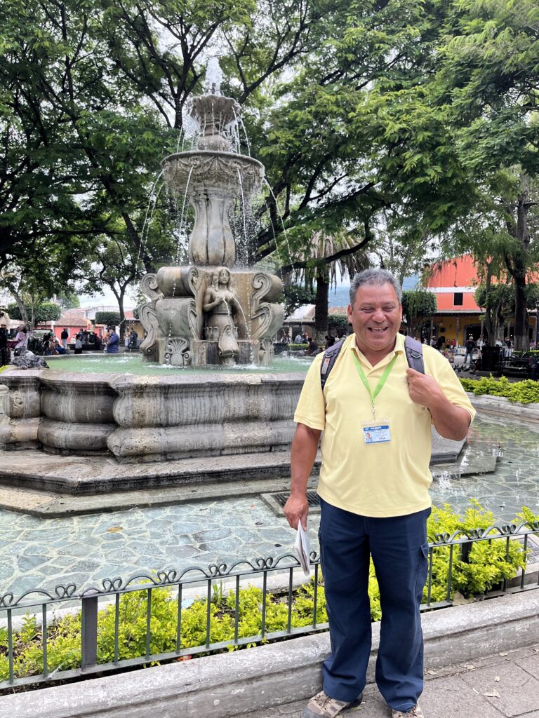 A man in a bright yellow shirt stands in front of a large stone fountain.