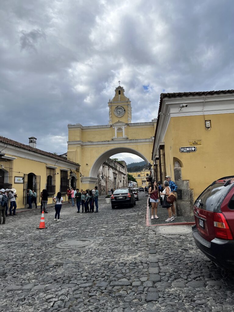 A yellow and white arch with a clock tower at the top spans a cobblestone street. Cars drive beneath it, and groups of tourists stop to take photos.