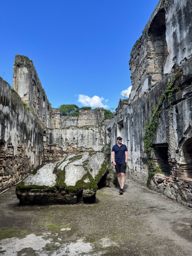 A man in a cap walks between mossy cement blocks that are bigger than he is. Around him are crumbling masonry walls with plants growing from them.