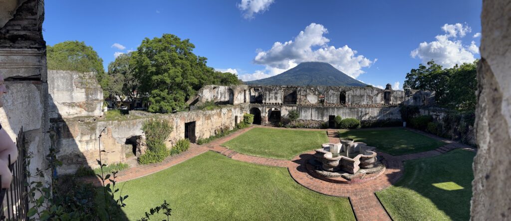 A large grassy courtyard with a stone fountain in the center and brick pathways throughout. The courtyard is surrounded by crumbling stone walls, and in the background is a pointed volcanic mountain, covered in trees, with clouds obscuring its peak.